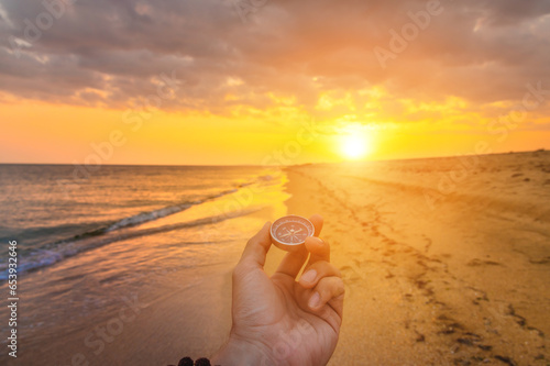 First person view of a man's hand with a compass against the backdrop of a beautiful seascape. Navigation concept of finding your way and orientation. The setting sun with rays enters the frame