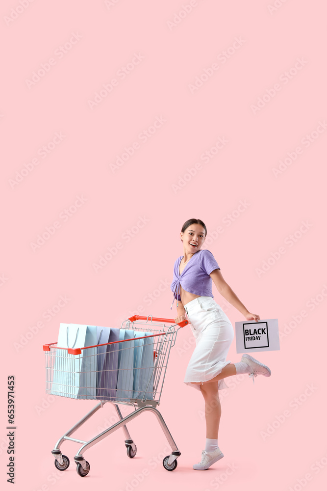 Young Asian woman with shopping bags in cart on pink background. Black Friday sale Young Asian woman with shopping bags in cart on pink background. Black Friday sale
