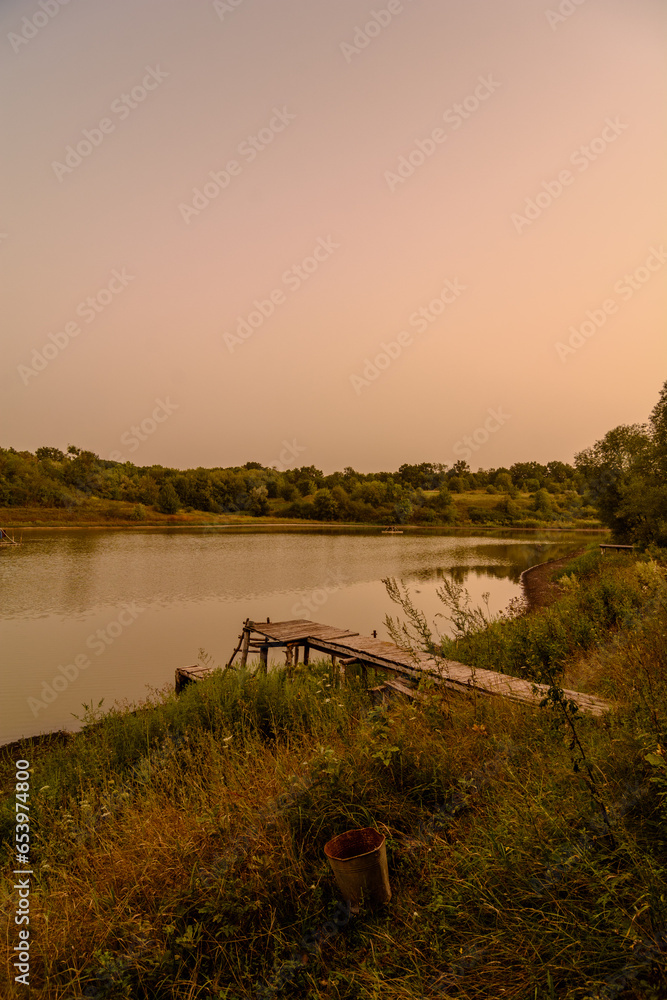 Wooden fishing pier over a small lake, against a backdrop of warm orange sky.
