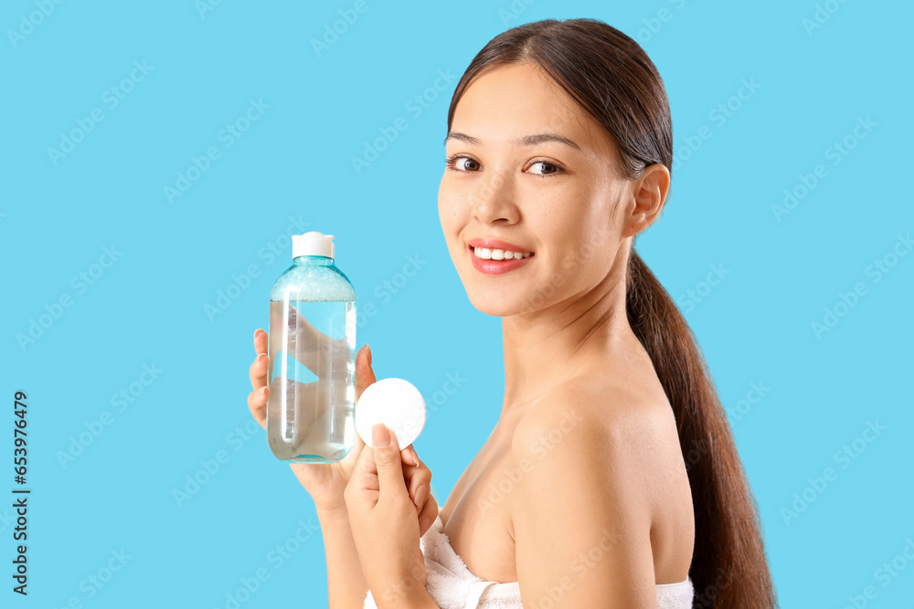 Young Asian woman with bottle of micellar water and cotton pad on blue background, closeup Young Asian woman with bottle of micellar water and cotton pad on blue background, closeup