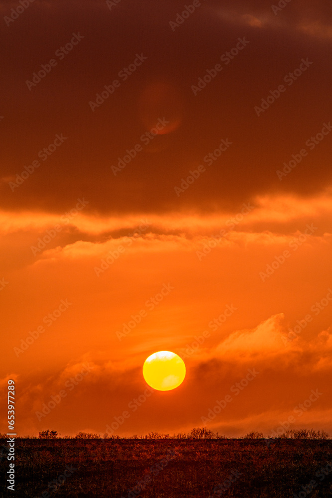 Bright orange sun close to the horizon against a cloudy sky during sunset, with an empty field in the foreground Bright orange sun close to the horizon against a cloudy sky during sunset, with an empty field in the foreground
