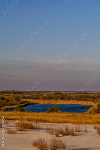 Landscape of a large square-shaped blue lake surrounded by yellow reeds and trees  with white sand in the foreground and a blue sky with clouds in the background.