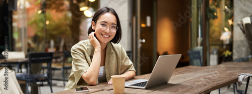 Portrait of smiling girl in glasses, sitting with laptop in outdoor cafe, drinking coffee and working remotely, studying online © Mix and Match Studio