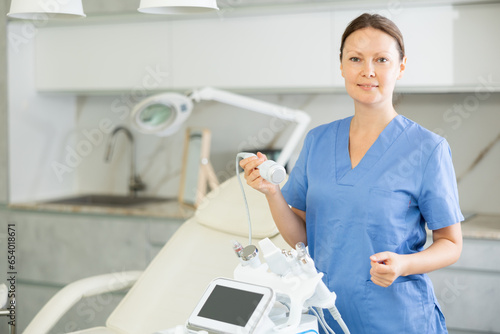 Adult female cosmetologist in medical uniform posing with cosmetology apparatus in medical room