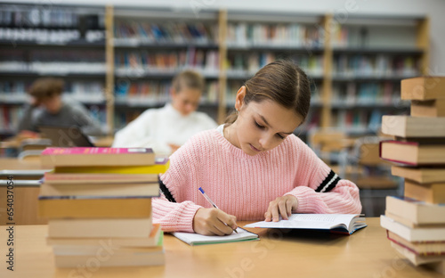Ten-year-old schoolgirl, preparing for lessons in the school library, takes notes in an exercise book of the necessary ..material from the textbook