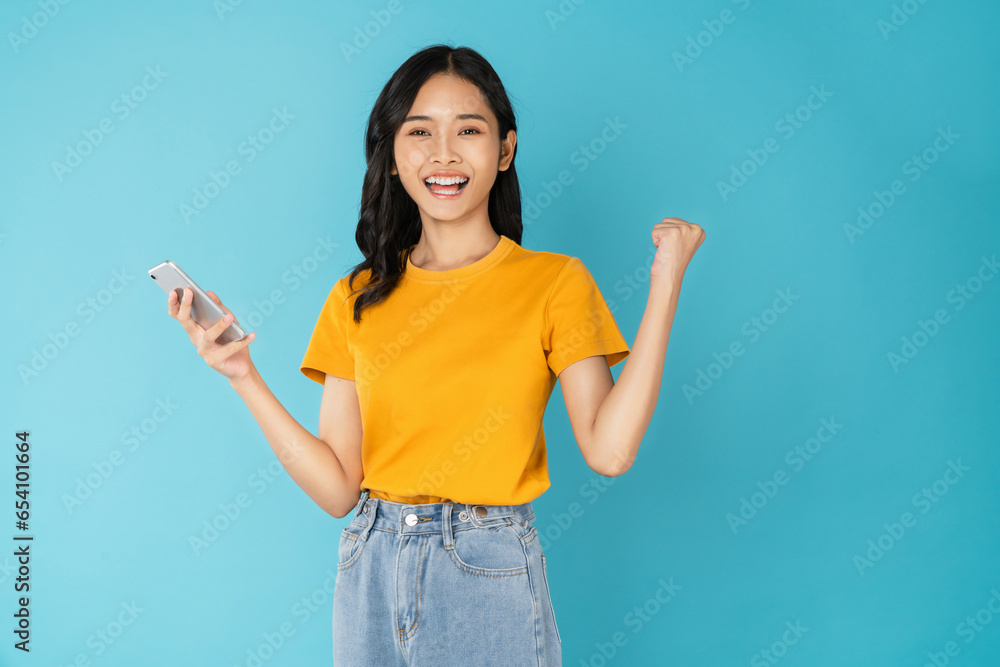Happy young Asian woman holding smartphone with fist up raise win on blue background. Happy young Asian woman holding smartphone with fist up raise win on blue background.
