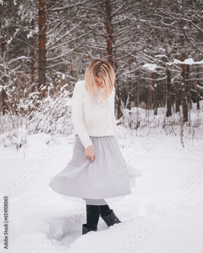 A young blonde girl wearing a white sweater is swirling her skirt in a winter snowy forest