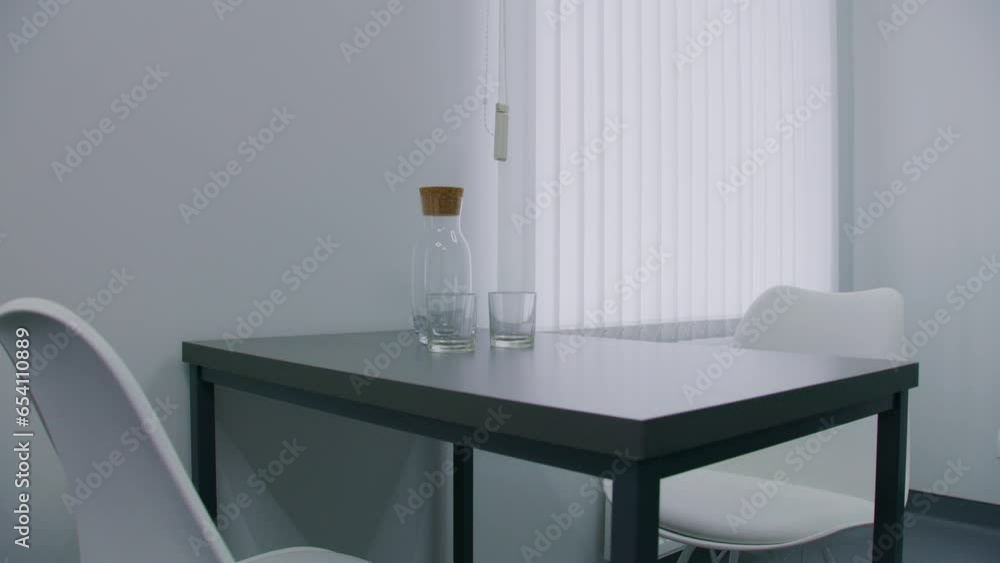 Close up of black dinner table with white chairs in middle of light hospital room near window. Glass bottle with glasses on table. Modern and comfortable hospital ward prepared for new patients. Close up of black dinner table with white chairs in middle of light hospital room near window. Glass bottle with glasses on table. Modern and comfortable hospital ward prepared for new patients.