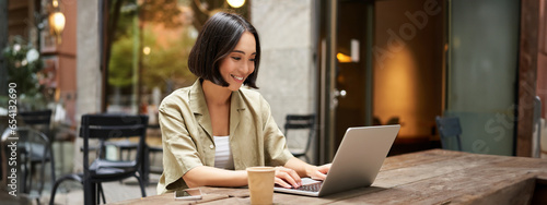 Young asian woman, digital nomad working remotely from a cafe, drinking coffee and using laptop, smiling