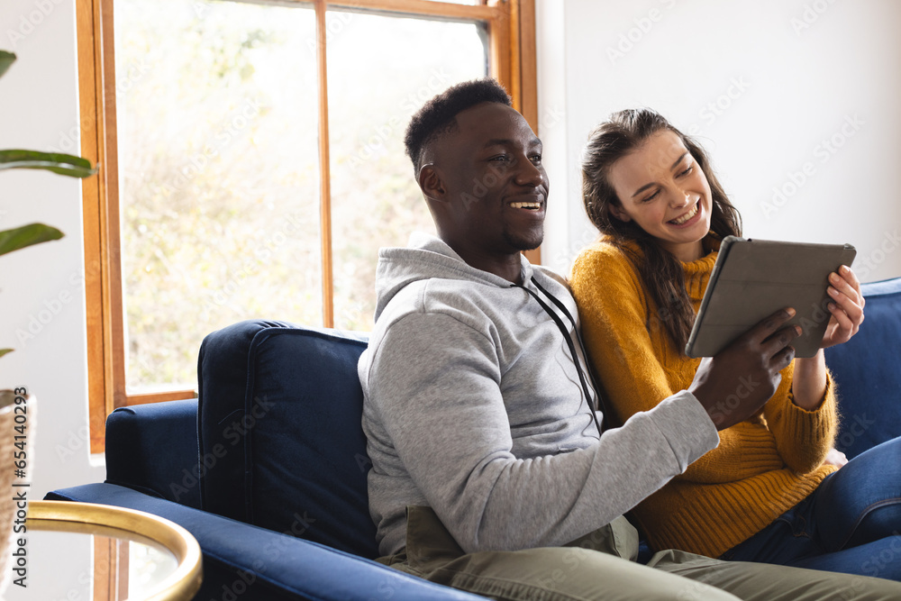 Happy diverse couple sitting on sofa using tablet at home, copy space