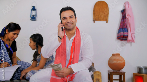 Indian villager taking a phone call about receiving money - financial concept. Village scene of a middle-aged farmer attending a phone call while counting his hard-earned income/salary
