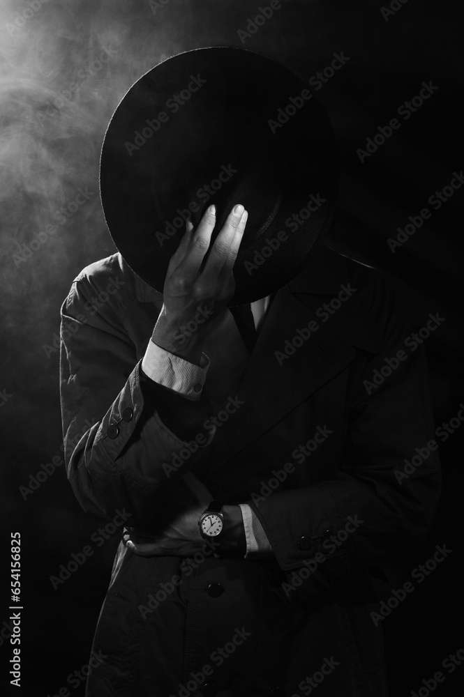 a dark silhouette of a man in a raincoat and a hat in the style of crime noir. Dramatic noir portrait in the style of detectives of the 1950s. There is smoke in the photo - obrazy, fototapety, plakaty 
