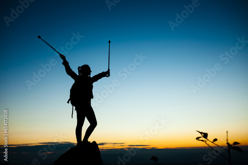 Silhouette of a hiker girl on a rock pedestal with hands up. Beautiful orange sunset on blue sky.