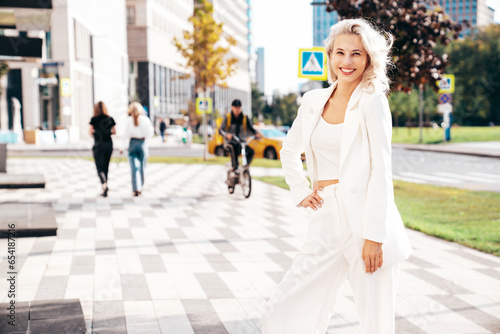Young beautiful blond woman wearing nice trendy white suit jacket. Smiling model posing in the street at sunny day. Fashionable female outdoors. Cheerful and happy