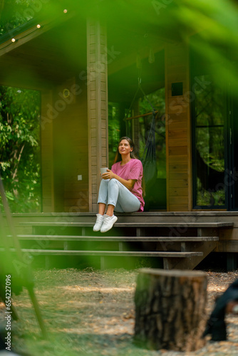 Konstfotografi A thoughtful girl sits with a cup of tea on the terrace of her wooden cottage in