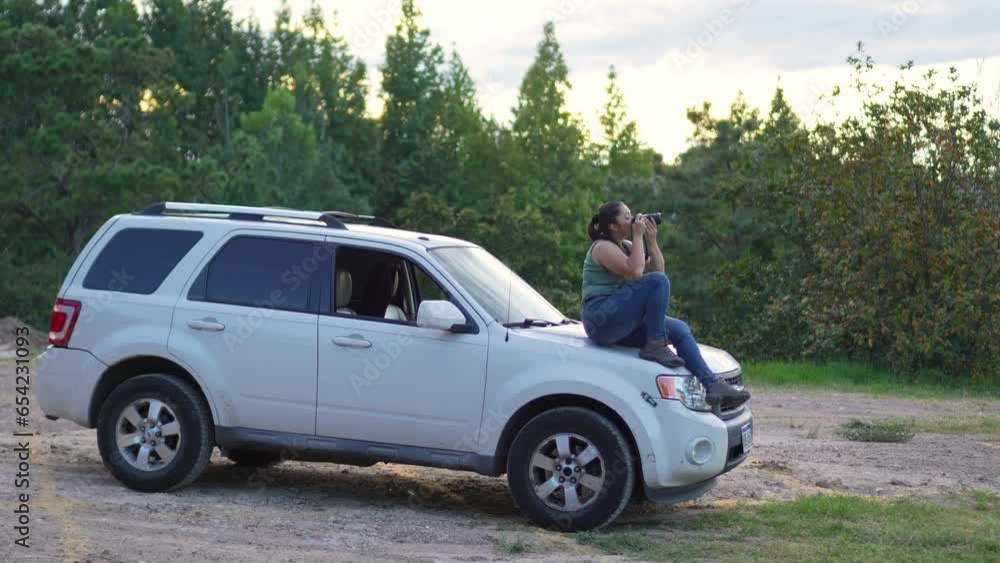 Woman with DSLR camera in her hands, sitting on the hood of her 4x4 SUV offroad car, taking photographs outdoors during sunset. - Long shot. Travel concept. Woman with DSLR camera in her hands, sitting on the hood of her 4x4 SUV offroad car, taking photographs outdoors during sunset. - Long shot. Travel concept.