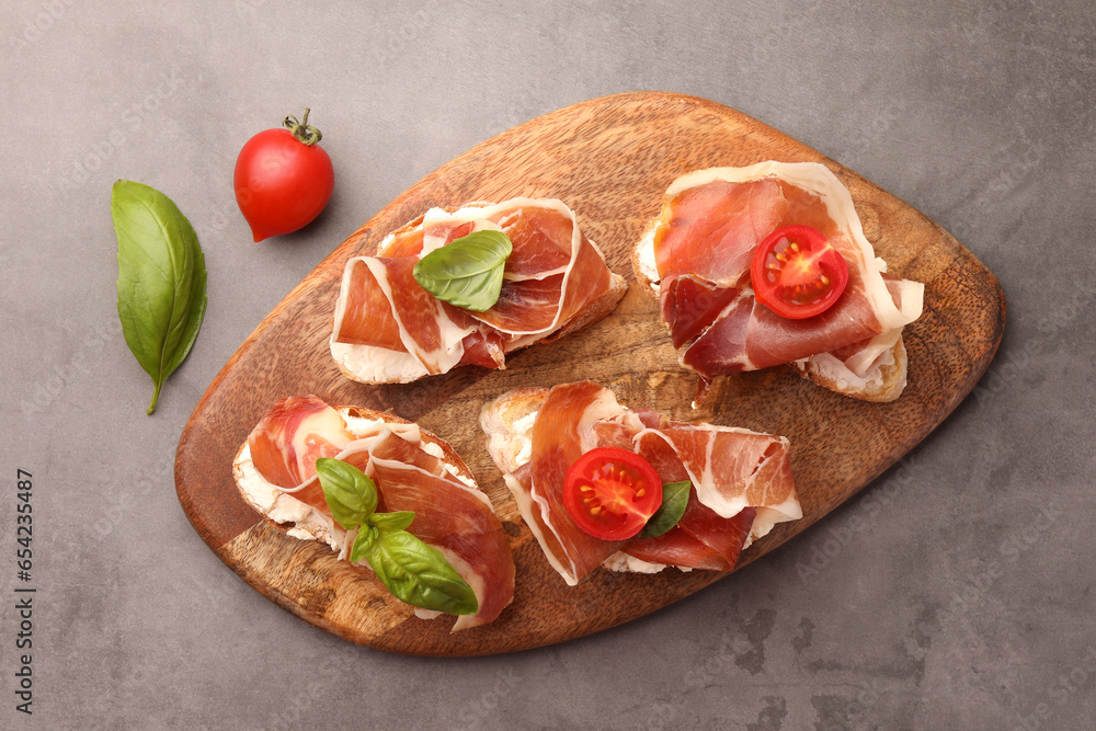 Tasty sandwiches with cured ham, basil and tomatoes on grey textured table, flat lay