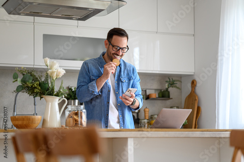Happy young businessman eating cookie while checking messages over mobile phone at kitchen counter