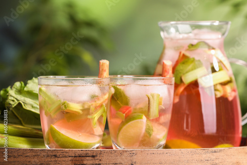 Glasses and jug of tasty rhubarb cocktail with citrus fruits on wooden board outdoors, closeup