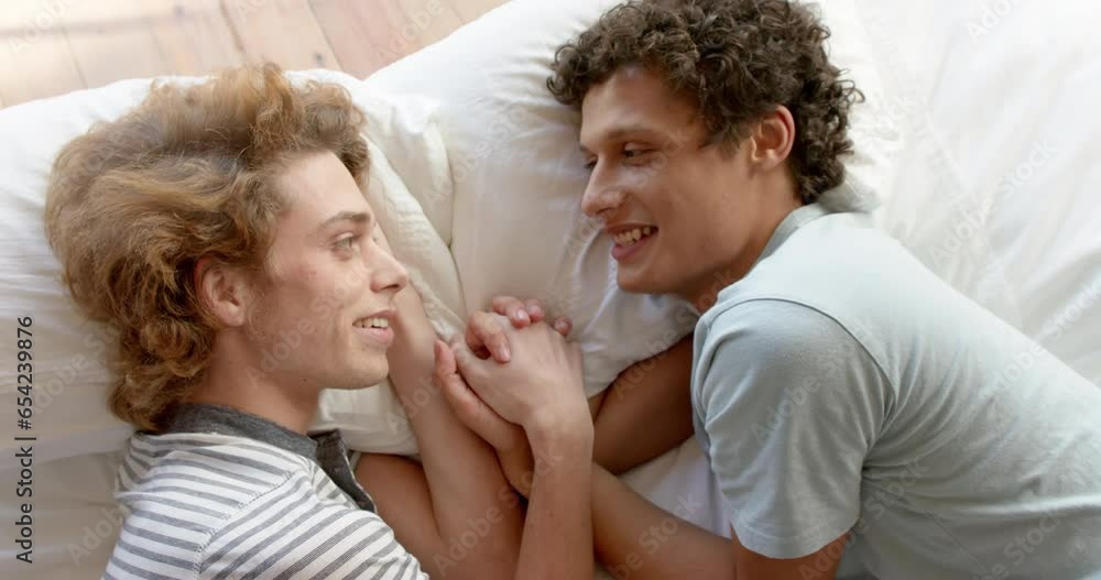 Happy diverse gay male couple lying on bed holding hands at home, slow motion Happy diverse gay male couple lying on bed holding hands at home, slow motion