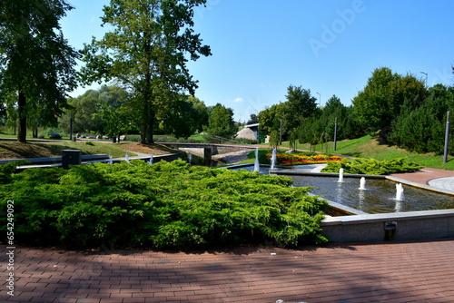 A close up on a well maintained park full of benches, fountains, shrubs, flower pots, forested areas and marble pavements seen on a sunny summer day near a small lake or river flowing through the area