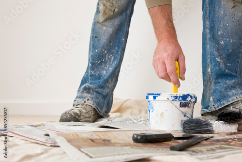 Close up of man legs wearing shoes and jeans covered in paint with his hand dipping a  paint brush
