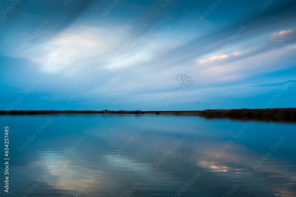 Paisaje azul de un lago al anochecer con una bandada de pajaros en el cielo.