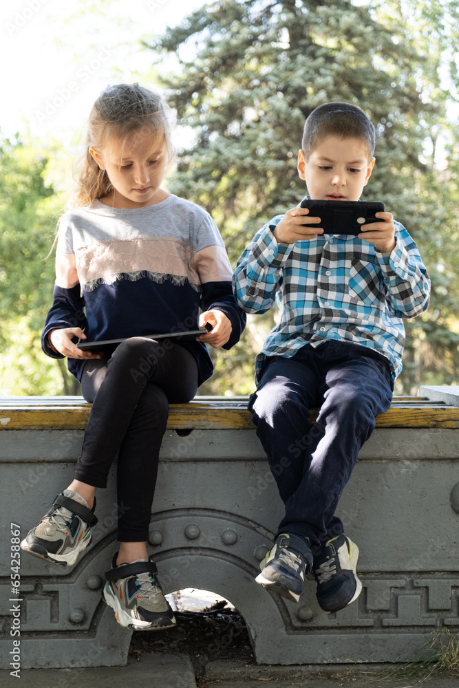 Children use gadgets while sitting in the park outdoors, games on smartphone and tablet