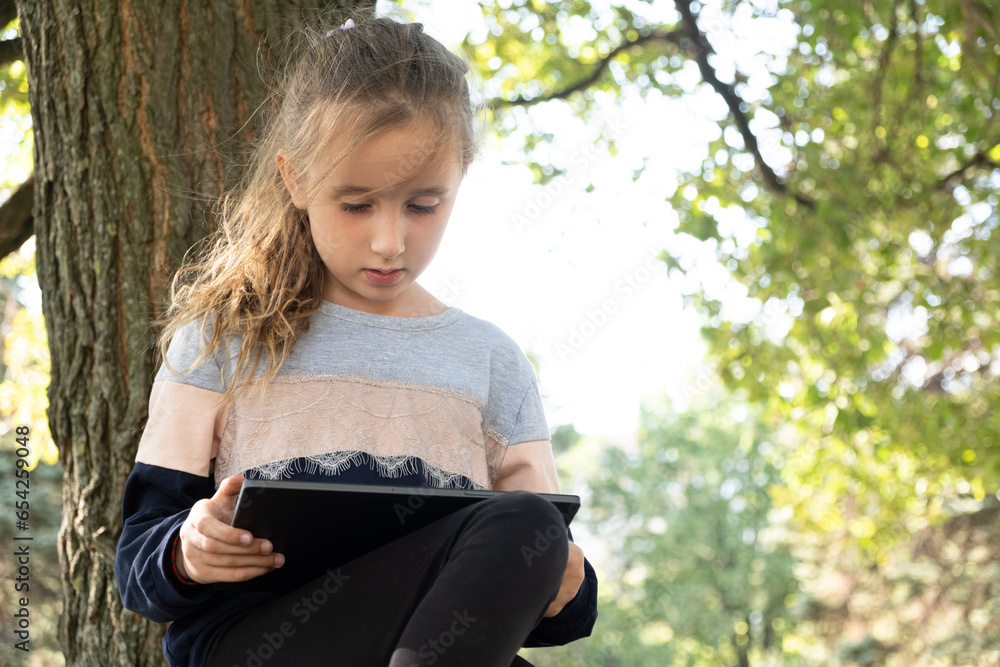 Little girl uses a tablet while sitting in the park in the summer. Little girl uses a tablet while sitting in the park in the summer.