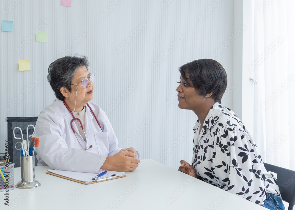 Asian senior elderly 60s physician wearing lab coat and stethoscope give advice to African patient woman in modern clinic, doctor visit with clients, working ability of aging people in special career