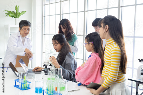 The ability of elderly employee working in science laboratory or education business, senior woman 60s with lab coat teaching group of Asian students looking microscope to analyze chemistry additives