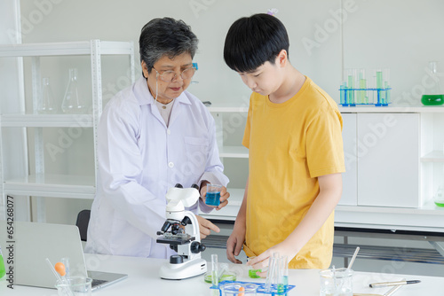 Asian cheerful schoolboy student using microscope standing with senior specialist teacher wearing lab coat in science laboratory  pupils learning by doing activities in class  elderly aging employment