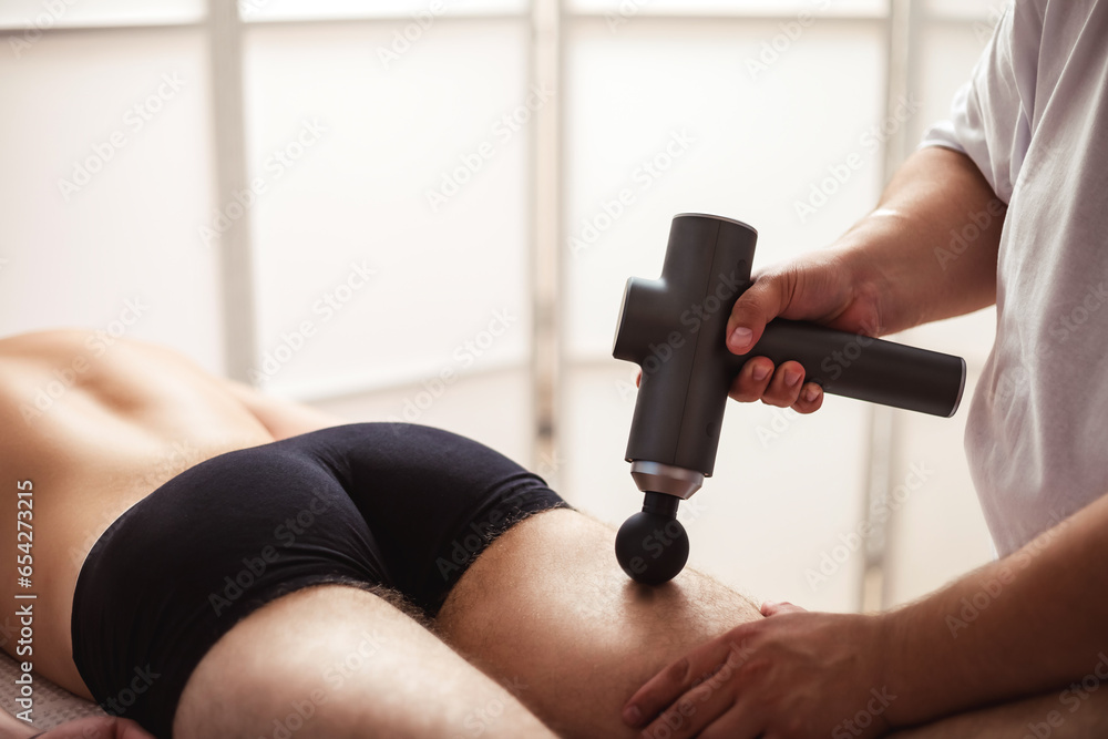 Doctor therapist treating injury of leg muscle sportsman of gun percussion massager. Masseur doing massage of thigh guy in medical clinic room. Alternative therapy rehabilitation. Copy ad text space - obrazy, fototapety, plakaty 