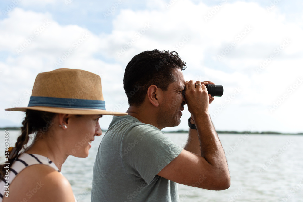 Obraz premium A man looks through a telescope at the horizon while a blurred woman wearing a hat looks in the same direction, with the ocean in the background.