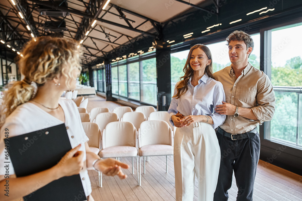 smiling couple in love looking at event organizer with clipboard talking in modern wedding venue smiling couple in love looking at event organizer with clipboard talking in modern wedding venue