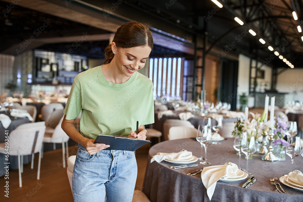 joyful event manager writing notes on clipboard near tables with
