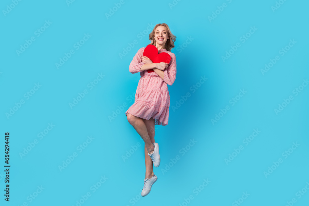 Full size photo of adorable cheerful girl dressed stylish pink clothes jumping cuddle red paper heart isolated on blue color background Full size photo of adorable cheerful girl dressed stylish pink clothes jumping cuddle red paper heart isolated on blue color background