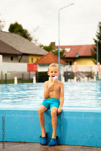 A seven-year-old boy of European appearance sits on the side of the pool and eats ice cream.