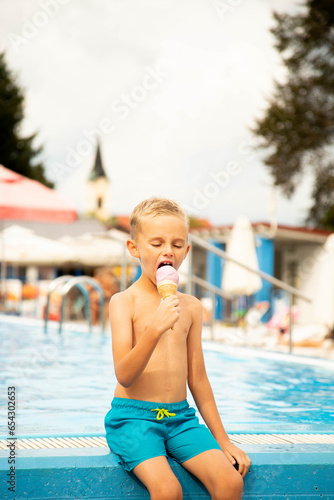 A seven-year-old boy of European appearance sits on the side of the pool and eats ice cream.