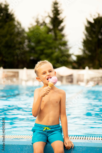 A seven-year-old boy of European appearance sits on the side of the pool and eats ice cream.