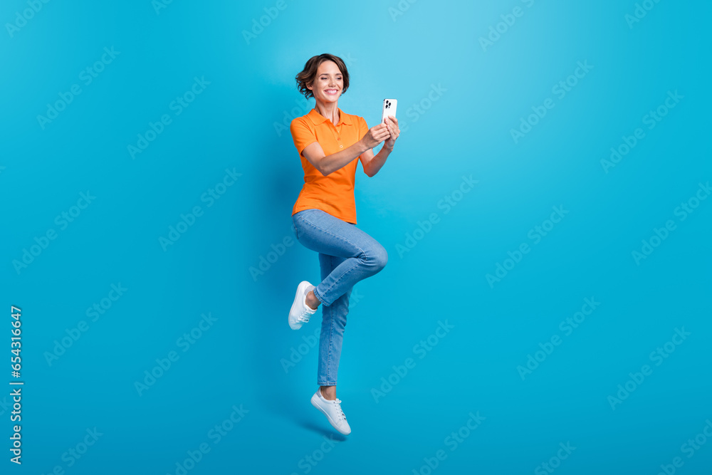 Full length photo of adorable optimistic woman wear orange t-shirt stylish jeans jumping make selfie isolated on blue color background