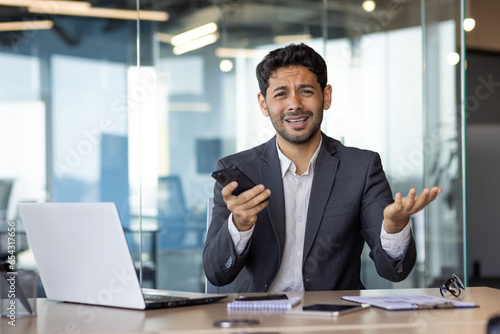 Problems in business negotiations, financial matters. A young Indian businessman is reading in the office at the table with the phone and is worriedly waving his hands.