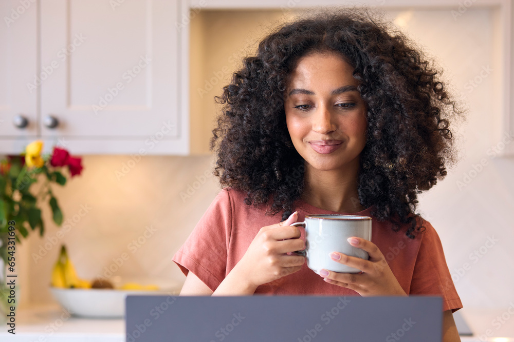 Young Woman In Kitchen At Home Looking At Laptop Holding Hot Drink Young Woman In Kitchen At Home Looking At Laptop Holding Hot Drink