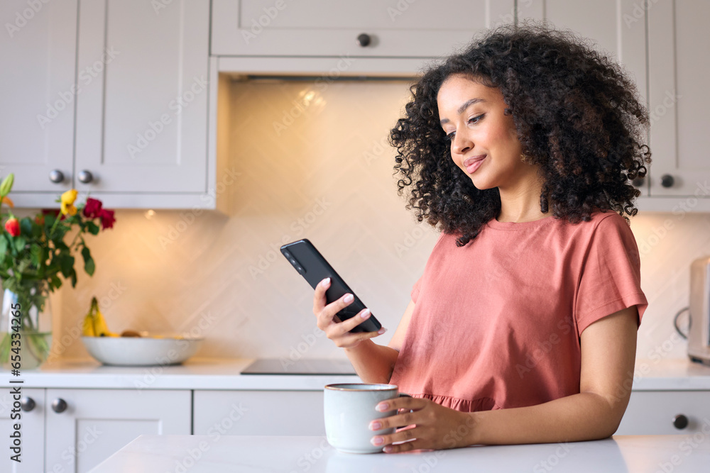 Young Woman Leaning On Kitchen Counter At Home Checking Social Media On Mobile Phone With Hot Drink Young Woman Leaning On Kitchen Counter At Home Checking Social Media On Mobile Phone With Hot Drink
