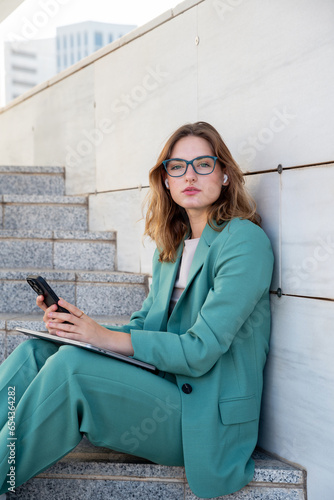 Business woman sitting on stairs outside office building looking at camera while using her earphones
