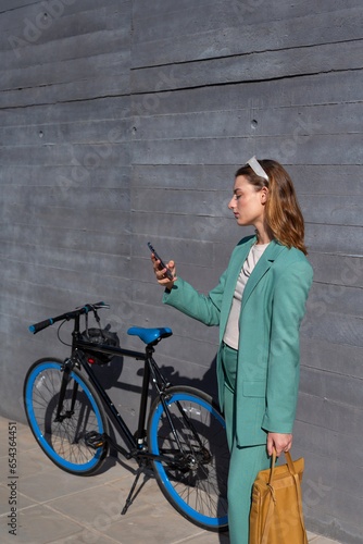 Young empowered ginger-haired woman in green suit using smartphone standing with her bike on black background