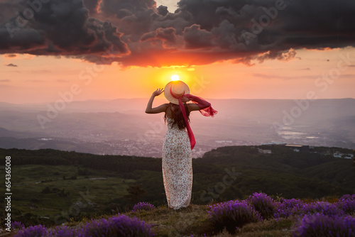 A young woman in a summer dress and a straw hat stands on a hill and looks at the setting sun over the city