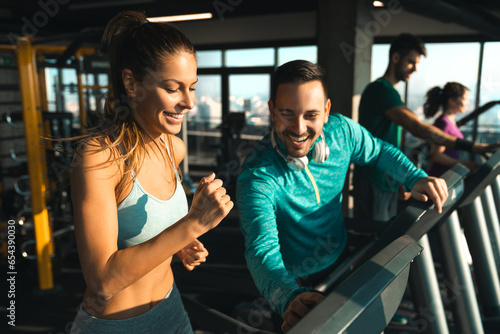 Young athlete male with headphones assisting a young woman while exercising on treadmills in a gym. Two people having engaging conversation during workout.