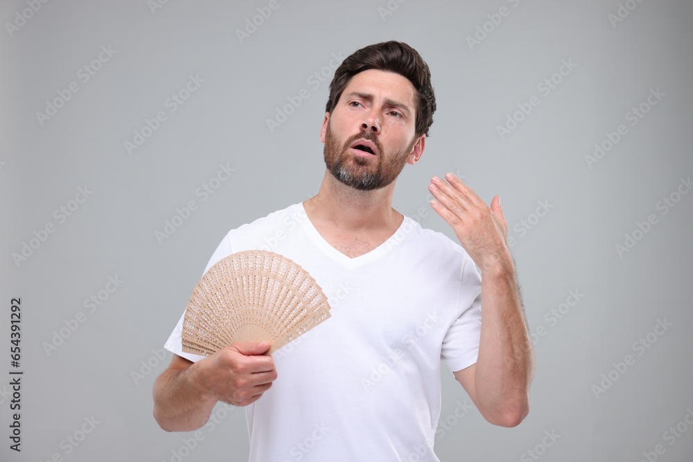 Unhappy man with hand fan suffering from heat on light grey background Unhappy man with hand fan suffering from heat on light grey background