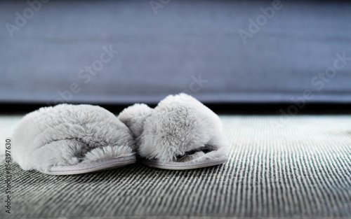 Gray fluffy house worn slippers stand on a melange-colored rug, near the sofa. The fur inside the slippers is crushed. Place for text. Home comfort.